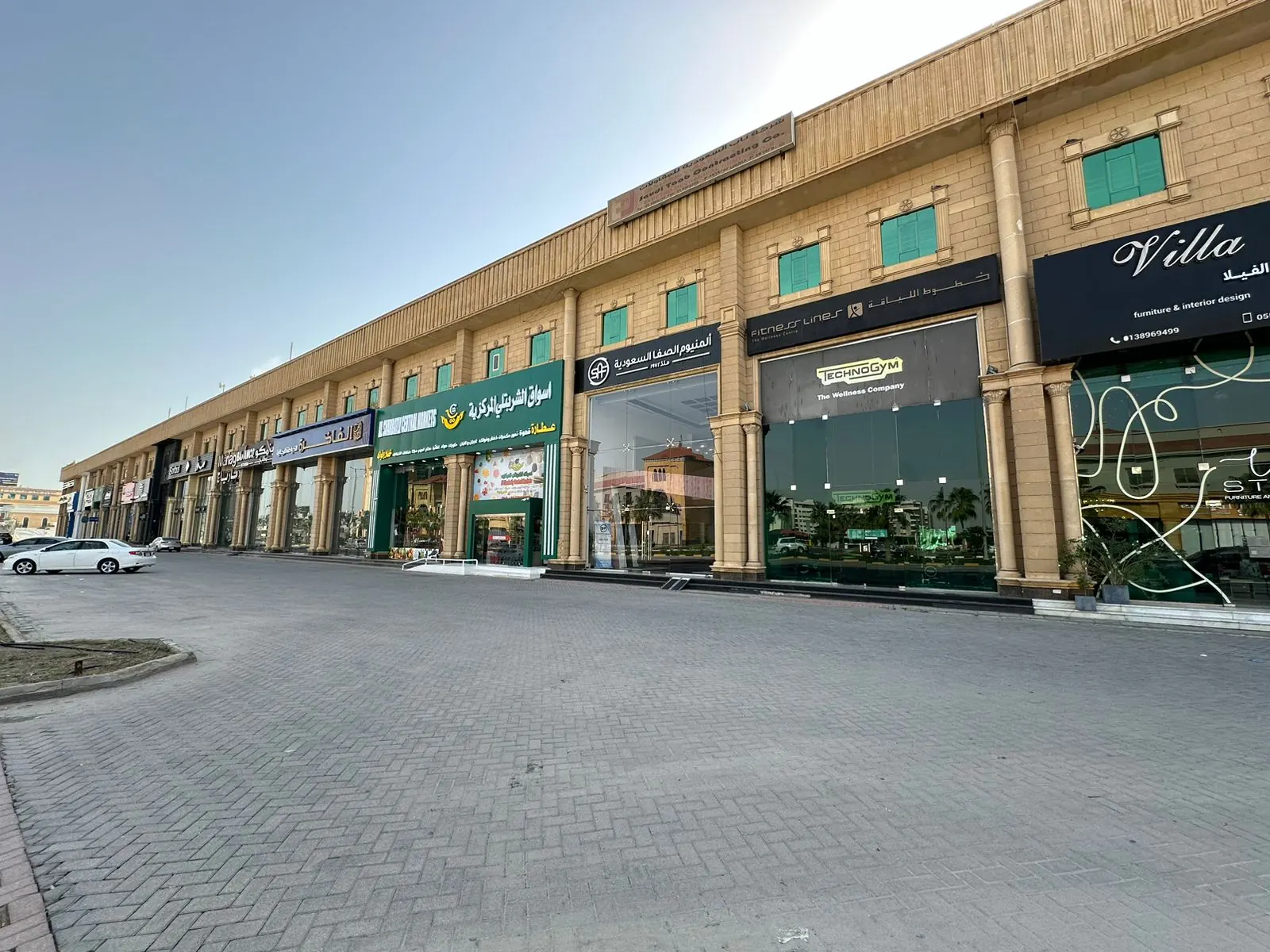 المجمع من ساحته المرصوفة: واجهة حجرية طويلة من مستويين، لافتات المعارض في الأسفل ونوافذ بمصاريع خضراء في الأعلى.
